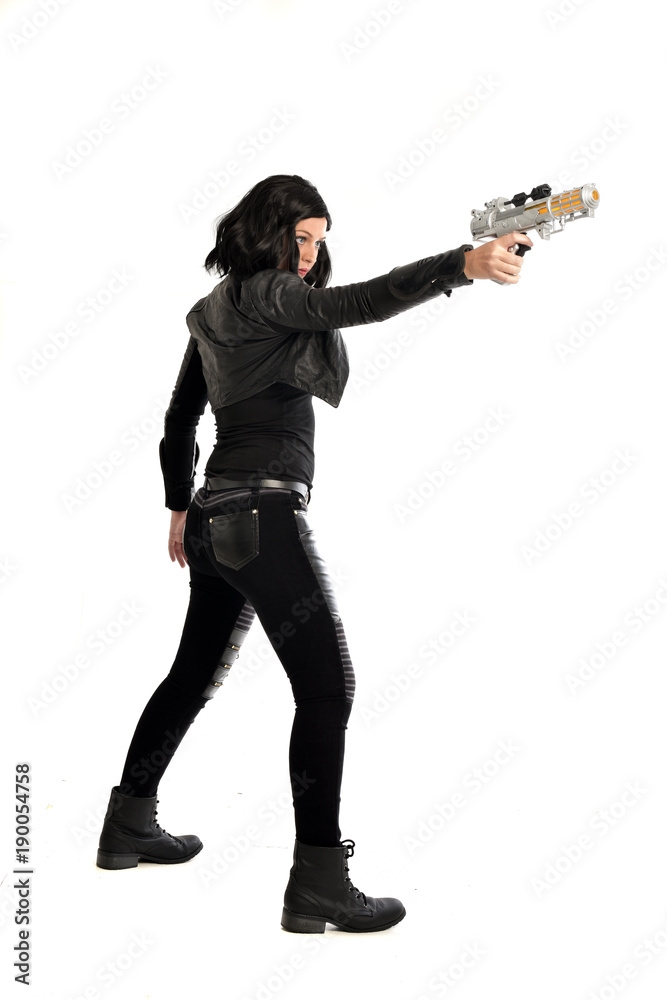 full length portrait of black haired girl wearing leather outfit. standing pose while holding a gun, isolated on a white studio background. full length portrait of black haired girl wearing leather outfit. standing pose while holding a gun, isolated on a white studio background.