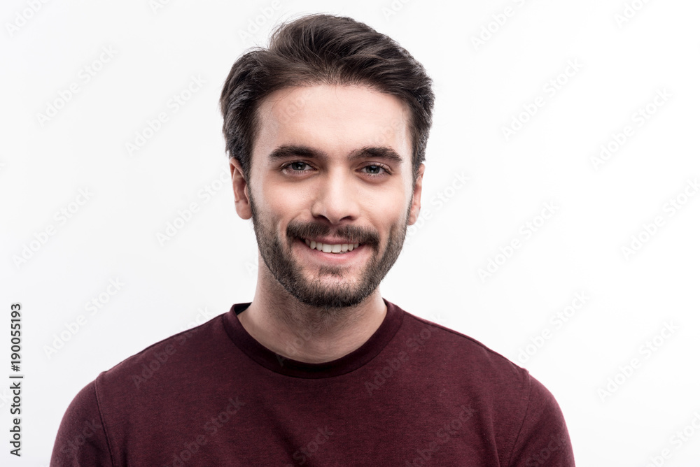 Epitome of masculinity. The portrait of a handsome dark-haired bristled young man in a burgundy sweater smiling at the camera while posing against a white background Epitome of masculinity. The portrait of a handsome dark-haired bristled young man in a burgundy sweater smiling at the camera while posing against a white background