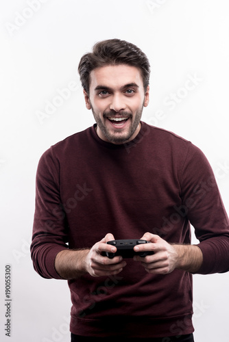 Game lover. Pleasant cheerful young man in a burgundy sweater holding a video game controller while posing against a white background © Viacheslav Yakobchuk