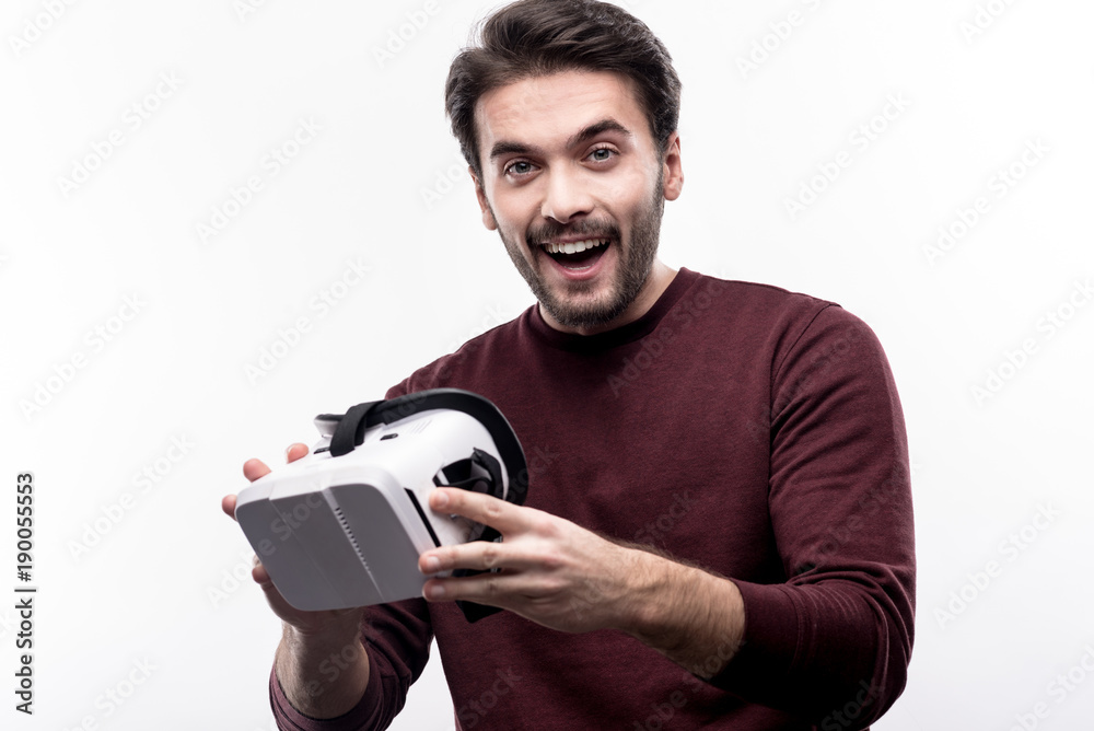 Happy about purchase. Charming upbeat young man in a burgundy sweater holding his new VR headset and smiling at the camera while standing isolated on a white background Happy about purchase. Charming upbeat young man in a burgundy sweater holding his new VR headset and smiling at the camera while standing isolated on a white background