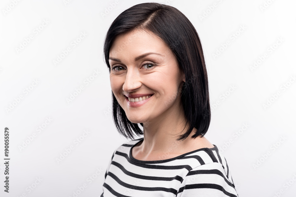 Charming female. The portrait of a pleasant joyful dark-haired woman in a striped pullover standing half-turned and posing for the camera with a smile Charming female. The portrait of a pleasant joyful dark-haired woman in a striped pullover standing half-turned and posing for the camera with a smile