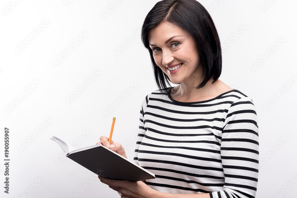 Planning thoroughly. Beautiful young woman in a striped pullover posing for the camera and smiling brightly while making notes in her planner Planning thoroughly. Beautiful young woman in a striped pullover posing for the camera and smiling brightly while making notes in her planner