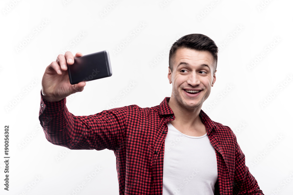 Nice shots. Pleasant young man in a checked shirt on top of a white t-shirt posing for the camera while taking selfies, using his phone Nice shots. Pleasant young man in a checked shirt on top of a white t-shirt posing for the camera while taking selfies, using his phone