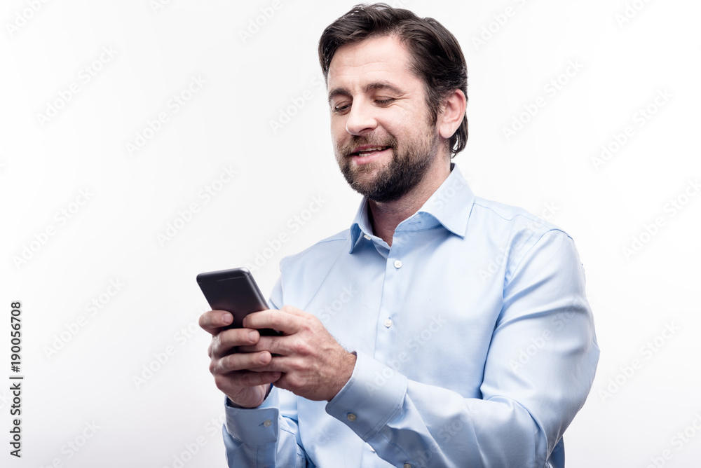 Pleasant chat. Cheerful middle-aged man in a baby blue shirt typing on his phone keyboard, sending messages to someone, while smiling fondly