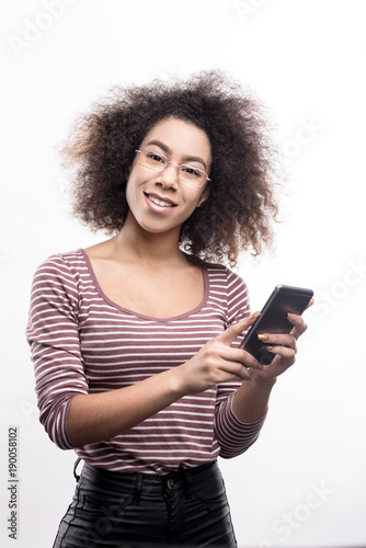 Connecting with friends. Adorable curly-haired young woman posing for the camera and smiling while holding her phone and sending text messages