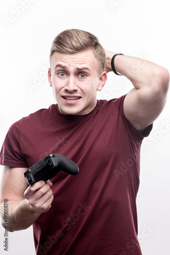 What a frustration. Handsome fair-haired young man in a burgundy shirt holding a game controller and clutching at his head in despair, having lost in a video game