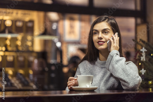 Young pretty brunette girl wears grey sweater have pleasant conversation by smartphone sitting near cafe window