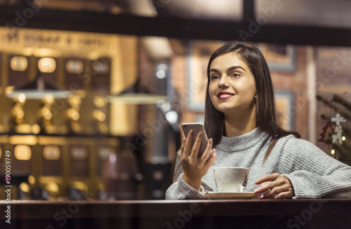 Young pretty brunette girl wears grey sweater holding smartphone sitting near cafe window