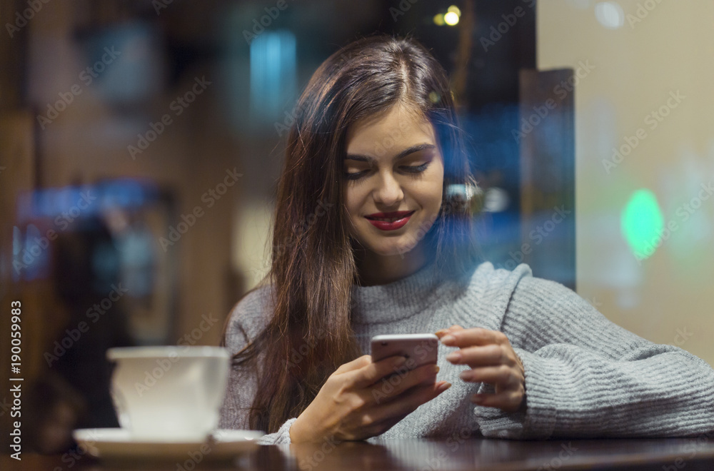Young pretty brunette girl wears grey sweater browsing internet via smartphone sitting near cafe window Young pretty brunette girl wears grey sweater browsing internet via smartphone sitting near cafe window