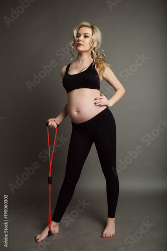 Pregnant beautiful blonde woman is wearing sportive black clothes doing exercises with rubber band isolated on gray, healthy life concept