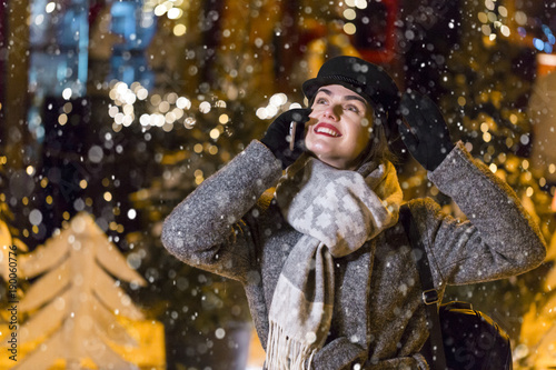 Stylish brunette smiling girl having pleasant talk by smartphone before garland wall in city snowy evening Christmas time