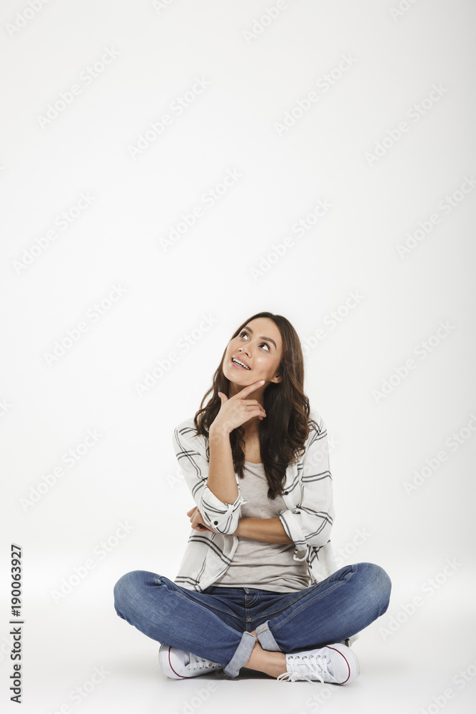 Full-length picture of brunette woman in casual clothes sitting in lotus pose on the floor with face upward and candid smile, isolated over white wall Full-length picture of brunette woman in casual clothes sitting in lotus pose on the floor with face upward and candid smile, isolated over white wall