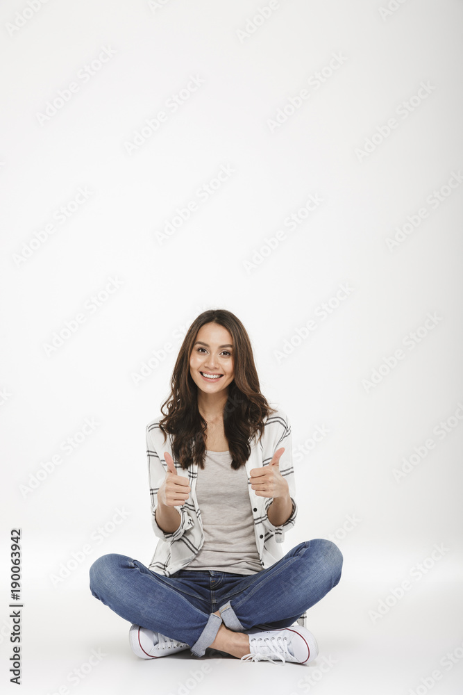 Full-length picture of smiling woman in jeans and sneakers sitting with legs crossed on the floor gesturing thumbs up, isolated over white wall Full-length picture of smiling woman in jeans and sneakers sitting with legs crossed on the floor gesturing thumbs up, isolated over white wall
