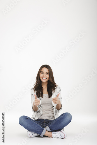 Full-length picture of smiling woman in jeans and sneakers sitting with legs crossed on the floor gesturing thumbs up, isolated over white wall