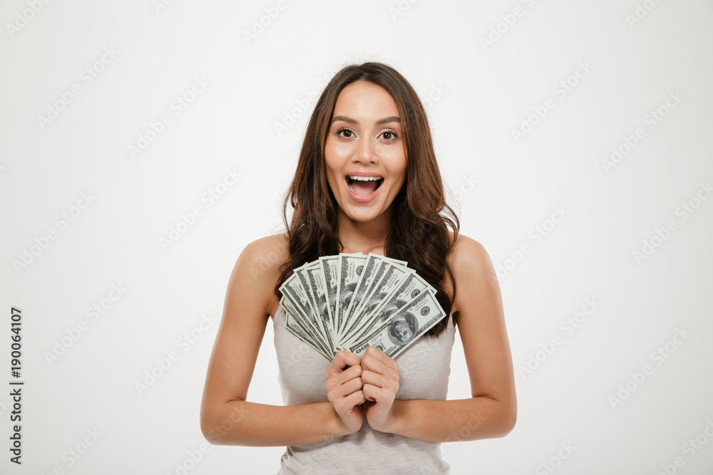Image of gorgeous brunette female model with long hair holding fan of 100 dollar bills, being rich and happy over white wall Image of gorgeous brunette female model with long hair holding fan of 100 dollar bills, being rich and happy over white wall