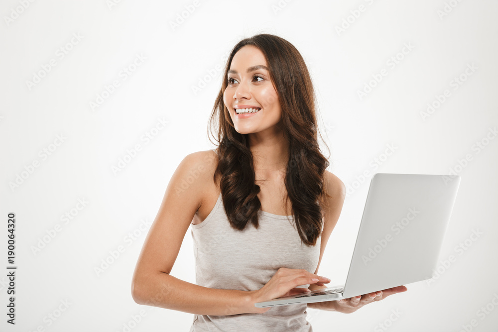 Photo of smiling woman with long brown hair looking away while working on silver personal computer, isolated over white wall Photo of smiling woman with long brown hair looking away while working on silver personal computer, isolated over white wall