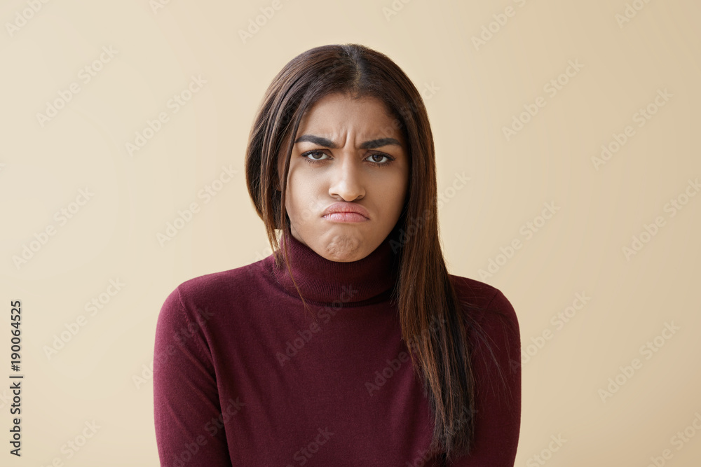 Fototapeta premium Portrait of upset young mixed race woman in maroon turtleneck pouting, feeling disappointed and unhappy after quarrel with her boyfriend, posing in studio with copyspace wall for your advertisement