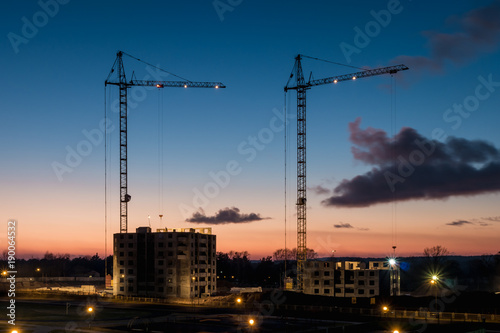 Tower cranes and unfinished multi-storey high near buildings under construction site in the sunset evening with dramatic colorful cloud background