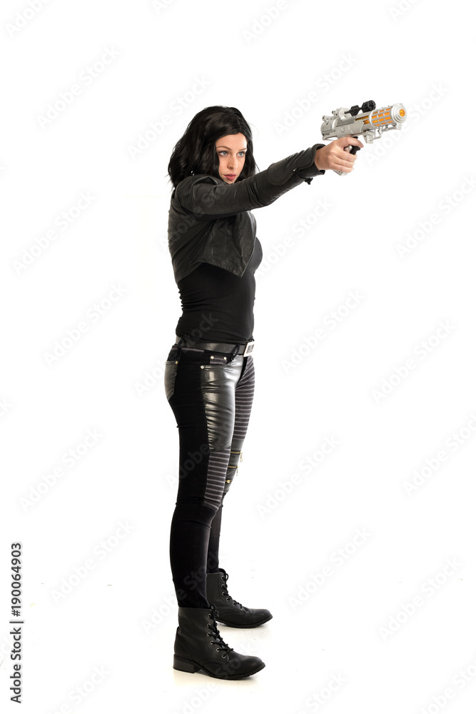 full length portrait of black haired girl wearing leather outfit. standing pose while holding a gun, isolated on a white studio background. full length portrait of black haired girl wearing leather outfit. standing pose while holding a gun, isolated on a white studio background.
