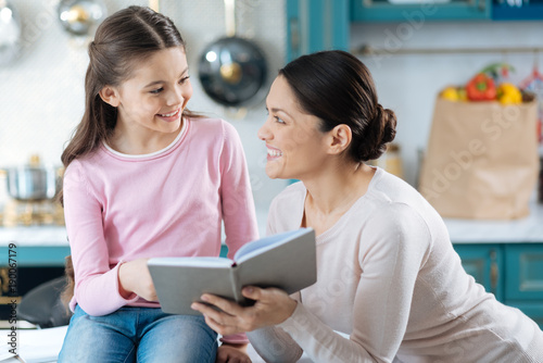 Happy family. Nice content dark-haired girl smiling to her mom while sitting on the table and her mum standing near her and holding a book