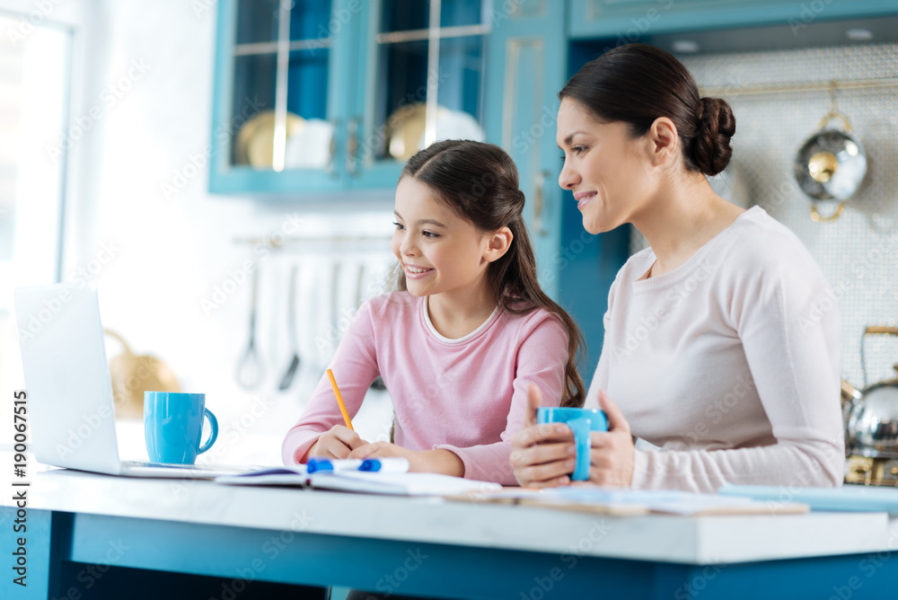 Learning lessons. Beautiful inspired dark-haired schoolgirl writing in her notebook and smiling while her mother standing near her with a cup Learning lessons. Beautiful inspired dark-haired schoolgirl writing in her notebook and smiling while her mother standing near her with a cup