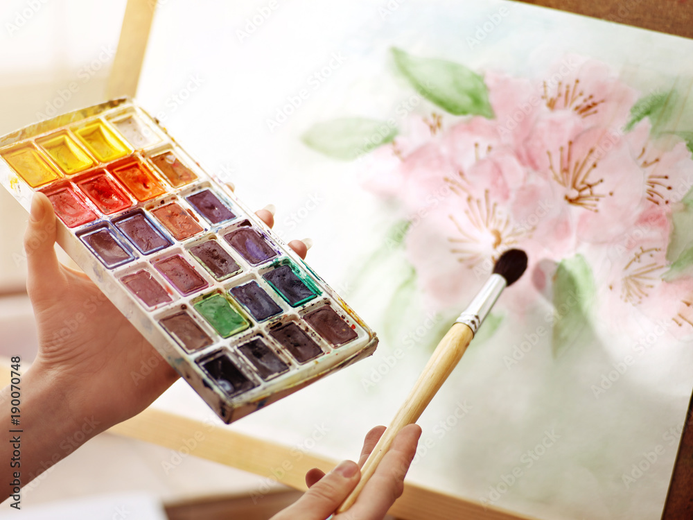 Obraz premium Artist painting on easel in studio. Authentic body part hands of girl paints with brush and set palette watercolor paints . Drawing of spring flowers.