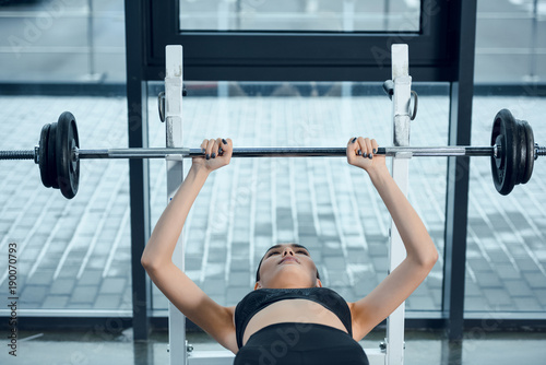 young fit woman lifting barbell while lying on bench at gym