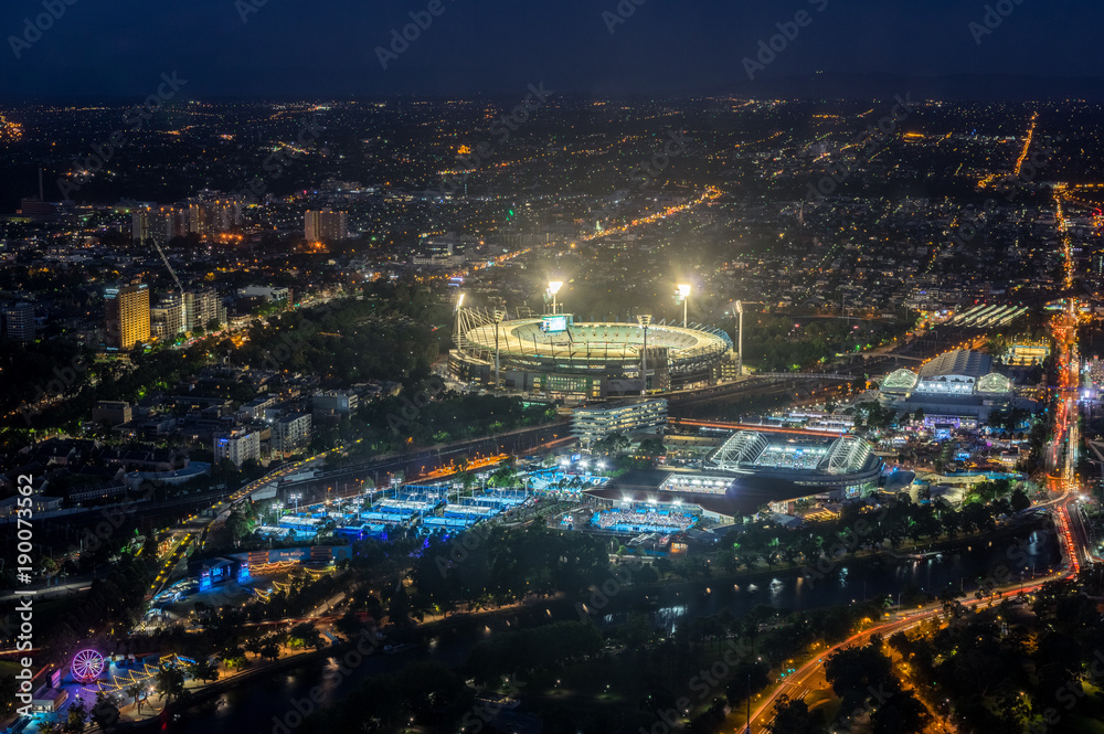 Melbourne Cricket Ground and Yarra Park tennis stadium illuminated at sunset.