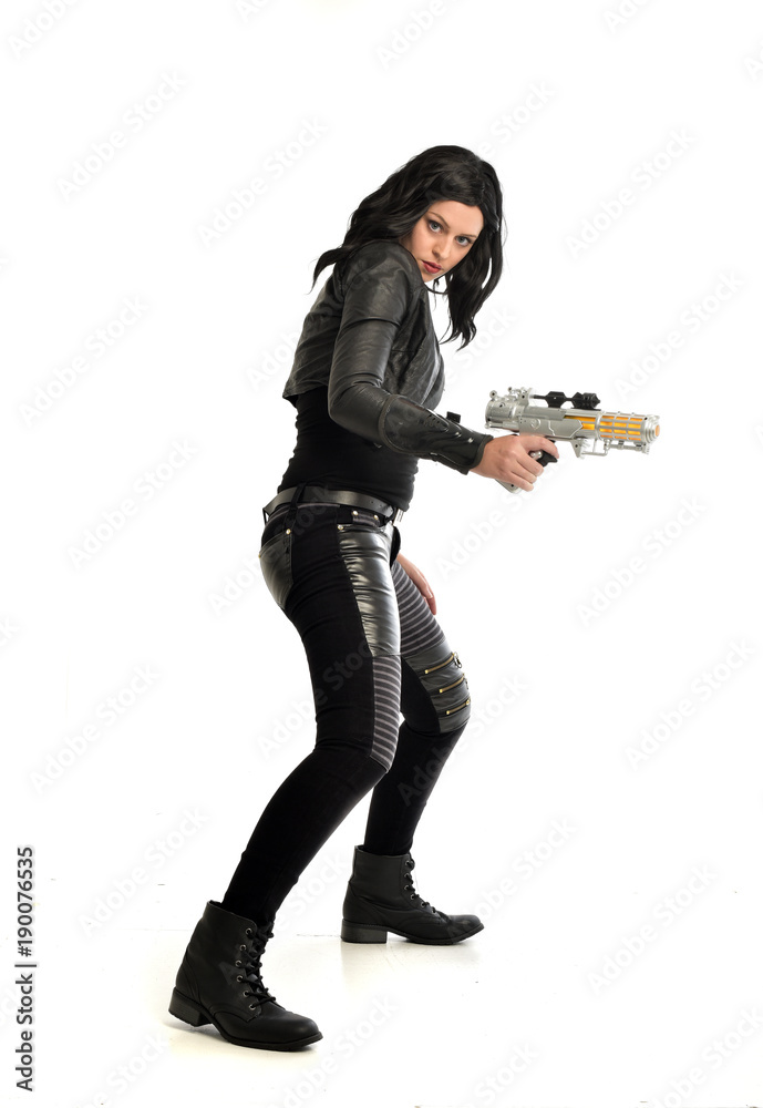 full length portrait of black haired girl wearing leather outfit. standing pose while holding a gun, isolated on a white studio background. full length portrait of black haired girl wearing leather outfit. standing pose while holding a gun, isolated on a white studio background.