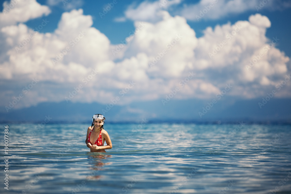 A young woman is standing in the sea against a background of clouds. She is dressed in a red swimsuit and on her head a mask for diving.
