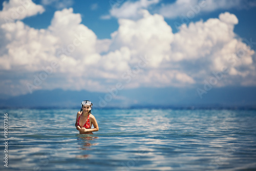A young woman is standing in the sea against a background of clouds. She is dressed in a red swimsuit and on her head a mask for diving.   © kazantsevaov