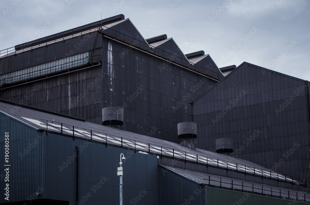 A group of dark grey factory buildings constructed from corrugated steel with a cctv security camera mounted on a pole A group of dark grey factory buildings constructed from corrugated steel with a cctv security camera mounted on a pole