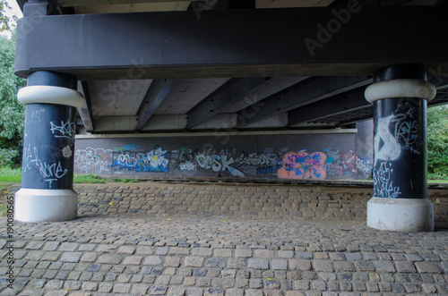 The underside of a steel road bridge which has had graffiti added to the support columns and brick wall