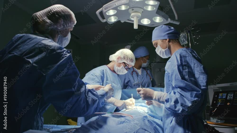 PAN of surgical team in scrubs and masks operating on patient in sterile room with modern equipment