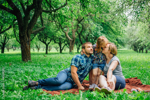 Young happy family of three lying on blanket in the park having fun. Happy parenting concept. Little girl kissing mother.