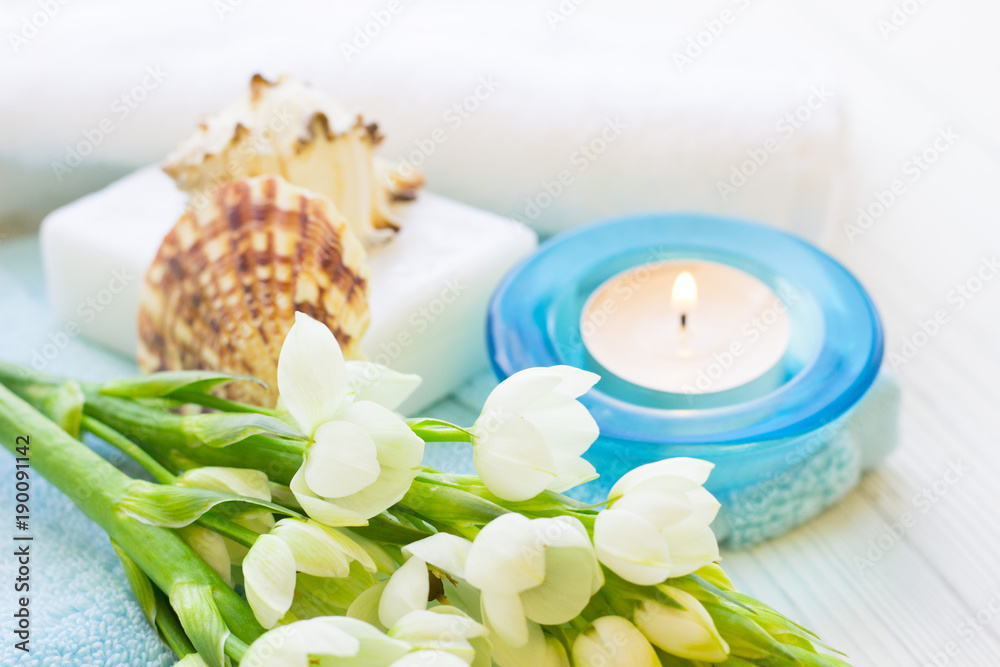 Aromatherapy Spa Concept with a fragrant candle in a blue candle holder, a bar of soap, terry towels, sea shells and white flower on white wooden background