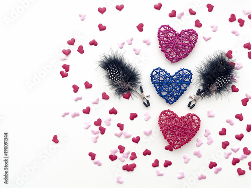 Three wicker hearts and nipple clamps with feathers (sex toy) are on a white background. Around these objects are scattered little figures of hearts. An image for Valentine's Day