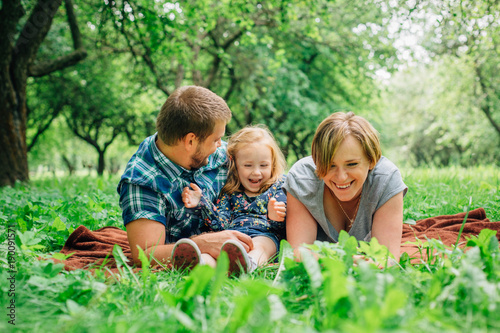 Young happy family of three lying on blanket in the park having fun. Happy parenting concept. Little girl with mother and father outdoors