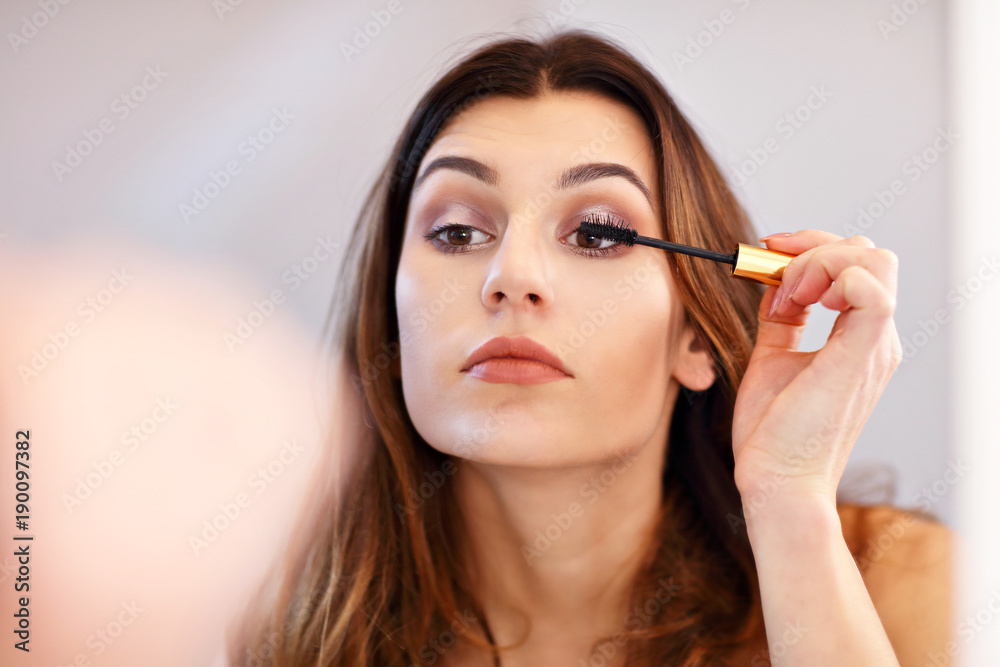 Attractive young woman doing make-up while looking at the mirror in bathroom