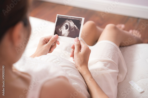 Pregnant asian woman looking at ultrasound scan of baby and touching her belly, close up of scan
