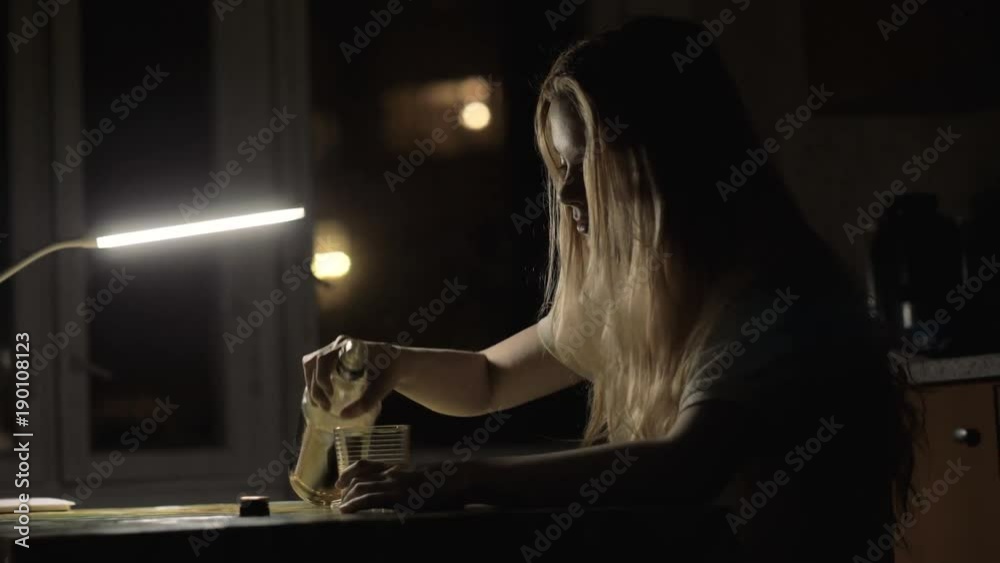 Young woman pouring alcohol in glass from a bottle and drinking in dark room