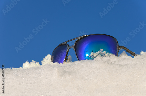 sunglasses in the shiny metal frame lying on the white snow under blue sky