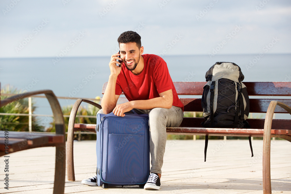 happy male traveler sitting on bench with bags and talking on mobile phone