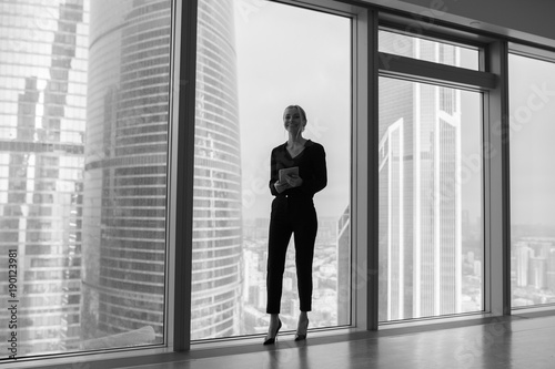 Business woman holding a tablet and standing in a modern office. Panoramic windows background. © kanashkin