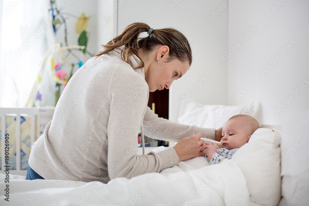 Cute sick child, baby boy, staying in bed, mom giving him medicine and checking on him