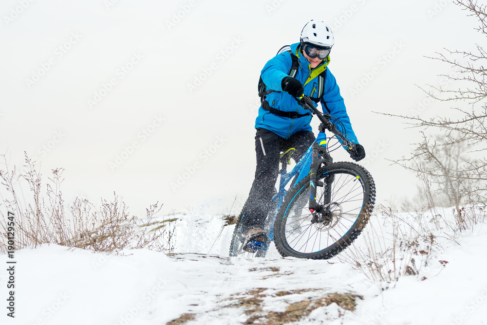 Cyclist in Blue Riding Mountain Bike on Rocky Winter Hill Covered with Snow. Extreme Sport and Enduro Biking Concept. Cyclist in Blue Riding Mountain Bike on Rocky Winter Hill Covered with Snow. Extreme Sport and Enduro Biking Concept.