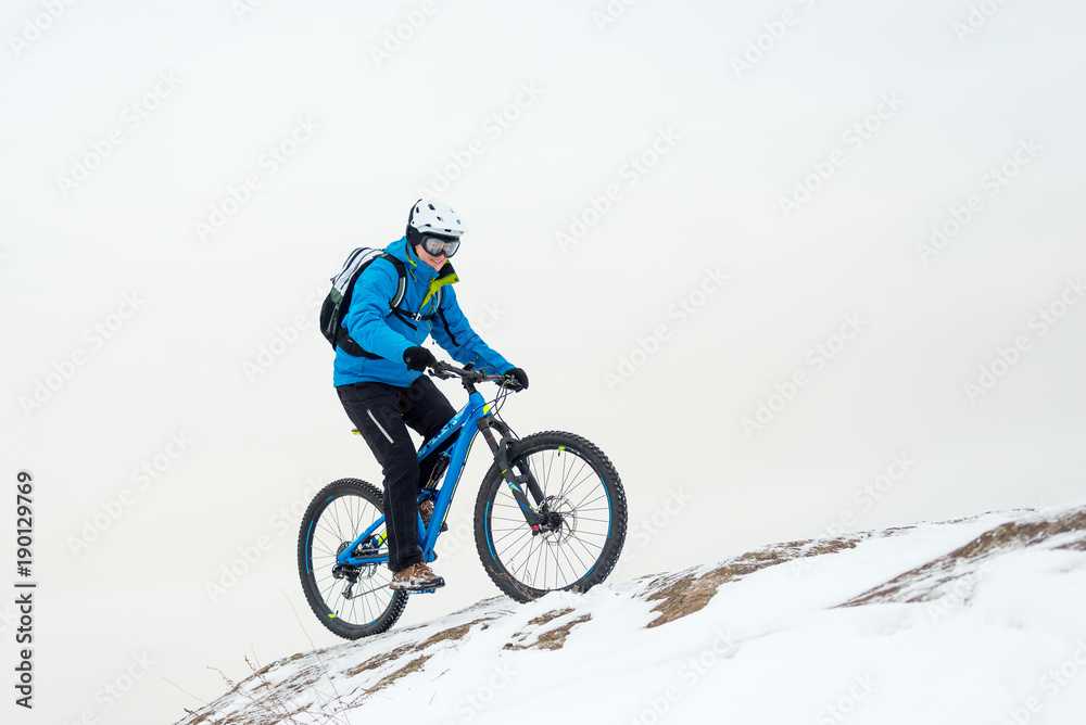 Cyclist in Blue Riding Mountain Bike on Rocky Winter Hill Covered with Snow. Extreme Sport and Enduro Biking Concept.