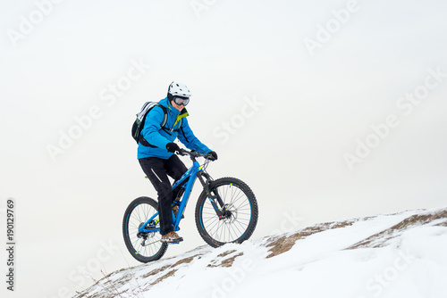 Fototapeta Naklejka Na Ścianę i Meble -  Cyclist in Blue Riding Mountain Bike on Rocky Winter Hill Covered with Snow. Extreme Sport and Enduro Biking Concept.