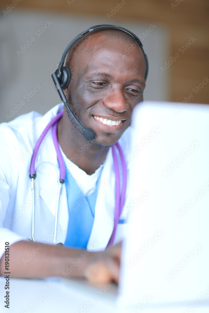 Portrait of young male doctor wearing headset while using computer at desk in clinic. Doctor. Portrait of young male doctor wearing headset while using computer at desk in clinic. Doctor.