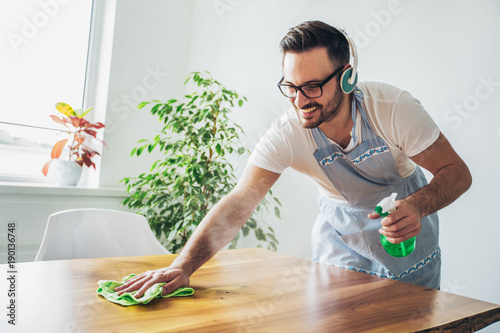 Young smiling man cleaning the table with a rag and spray bottle detergent